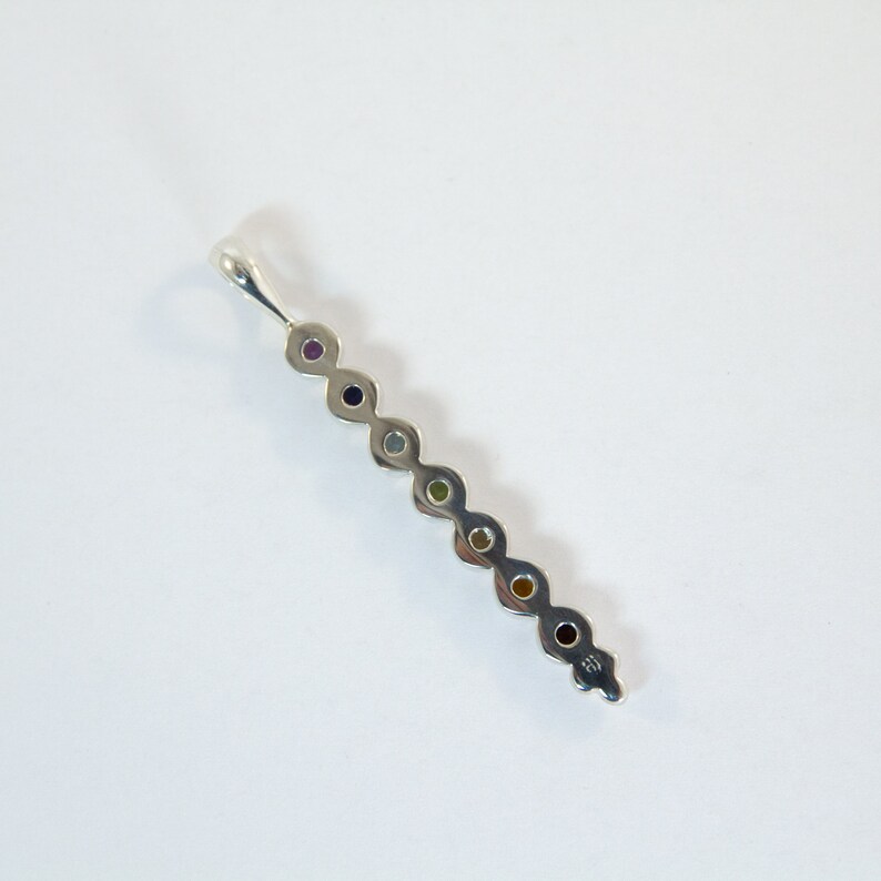 May include: A silver pendant featuring a linear design with seven colorful gemstones. The gemstones are arranged in a row, each a different color, including purple, blue, green, yellow, and red. The pendant has a loop at the top for a chain.