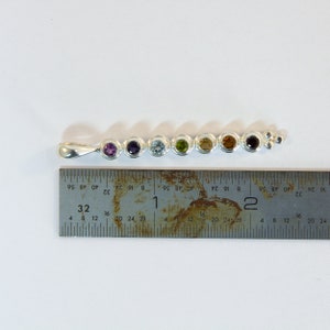 May include: A silver pendant featuring seven colorful gemstones in a row. The gemstones are purple, dark purple, light blue, green, yellow, orange, and dark red. A ruler is placed next to the pendant for scale.