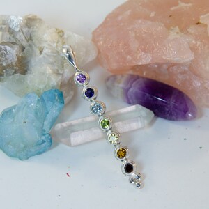 May include: A silver pendant featuring seven colorful gemstones in a row, representing the chakras. The gemstones are purple, indigo, light blue, green, yellow, orange, and red. The pendant is displayed with various crystals in the background.