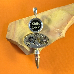 May include: A silver pendant featuring a round black button inscribed with "Shift Lock" and a silver oval revealing a clockwork mechanism. The pendant is attached to a silver bullet-shaped charm.
