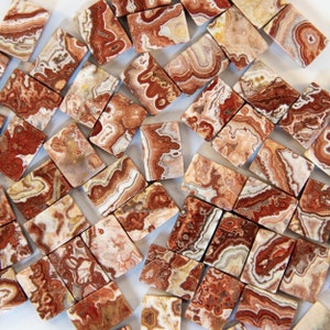 May include: A collection of small, rectangular, polished agate stones with a variety of brown, white, and red patterns. The stones are arranged in a random pattern on a white surface.