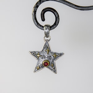 May include: A silver star-shaped pendant with a red gemstone in the center. The star is decorated with gold swirls and small stars.