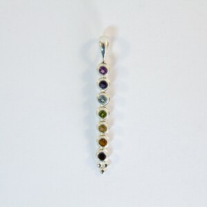 May include: A silver pendant featuring a vertical row of seven colorful gemstones. The gemstones are round and set in individual bezels, ranging in color from purple to red. The pendant has a loop at the top for a chain.