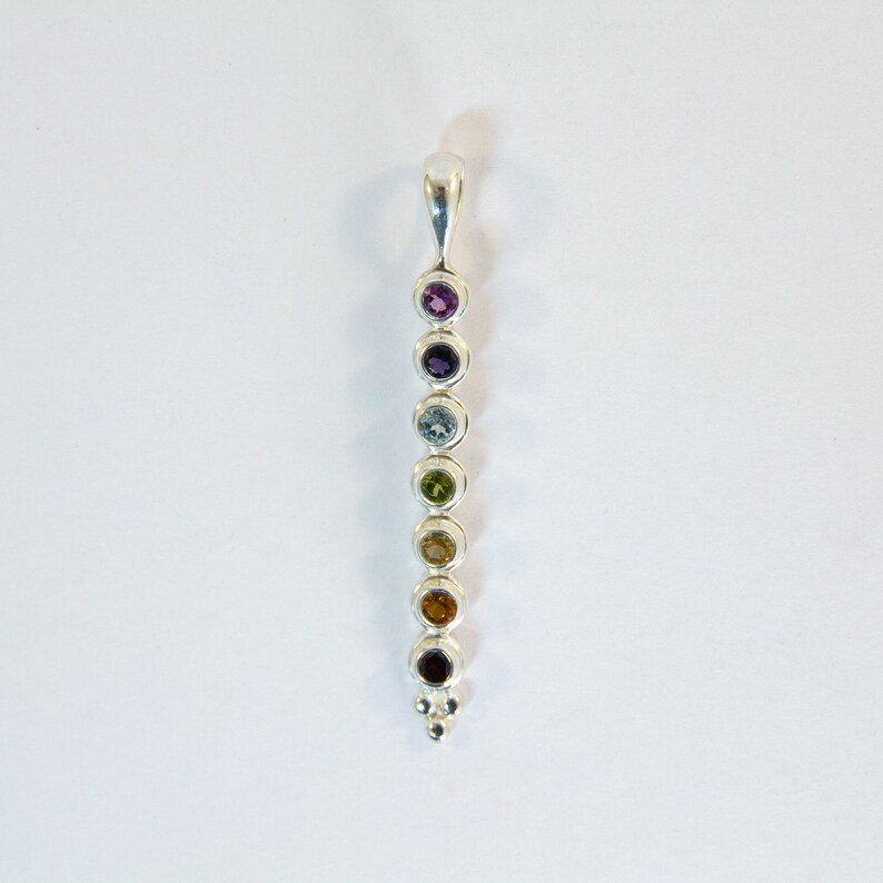 May include: A silver pendant featuring a vertical row of seven colorful gemstones. The gemstones are round and set in individual bezels, ranging in color from purple to red. The pendant has a loop at the top for a chain.