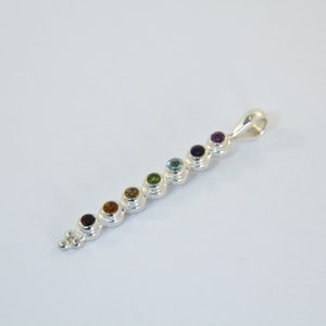 May include: A silver pendant featuring a series of seven colorful gemstones in a row. The gemstones are various colors, including red, orange, yellow, green, blue, and purple. The pendant has a loop at the top for a chain.
