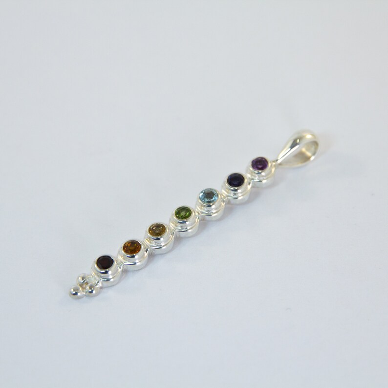 May include: A silver pendant featuring a series of seven colorful gemstones in a row. The gemstones are various colors, including red, orange, yellow, green, blue, and purple. The pendant has a loop at the top for a chain.