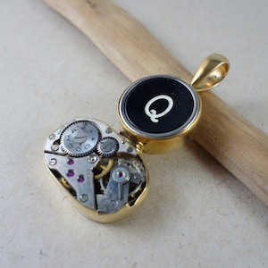 Puede incluir: Collar con colgante dorado con un dije de tecla de máquina de escribir con la letra 'Q' y un movimiento de reloj vintage en la parte posterior.