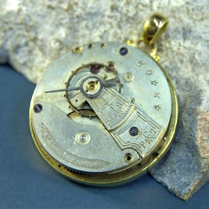 Puede incluir: Un colgante de reloj de bolsillo de oro con un mecanismo interno visible y la inscripción "MONTAUK WATCHES" en la parte posterior.