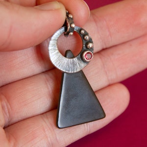 May include: A silver pendant with a black triangle and a circle with a red gemstone. The circle has a textured surface and a silver bail.