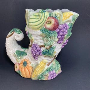 May include: A white ceramic cornucopia vase featuring a detailed design of grapes, corn, pumpkins, and apples. The vase is decorated with green leaves and a woven basket pattern.