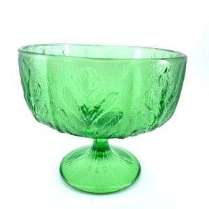 Puede incluir: Un cuenco verde de vidrio con pedestal, superficie texturizada y diseño de hojas. El cuenco tiene una parte superior ancha y abierta y una base redonda. La base tiene las letras "E.I.D." en relieve. El cuenco es probablemente vintage.