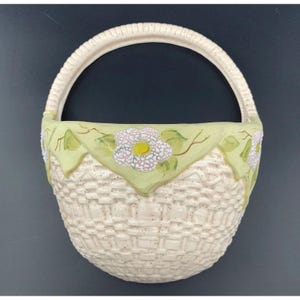 Puede incluir: Un bolsillo de pared de cerámica con forma de cesta tejida con una base de color crema y un acabado texturizado. El borde superior presenta un borde festoneado verde pálido decorado con flores blancas pintadas a mano, centros amarillos y tallos marrones.