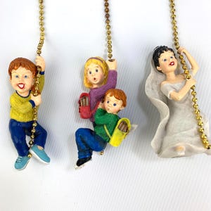Może przedstawiać: Trzy dekoracyjne figurki wiszące. Jedna to panna młoda w białej sukni i welonie. Pozostałe dwa to dzieci, jedno z żółtym swetrem i niebieskimi spodniami, a pozostałe dwa to dzieci z plecakami i swetrami. Wszystkie wiszą na złotym łańcuszku.