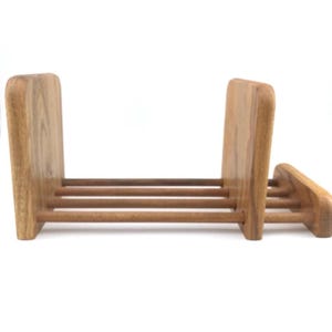 May include: A wooden bookend with a natural finish. The bookend has two upright rectangular supports connected by three horizontal dowels. A smaller, angled support is attached to the side. The wood grain is visible.