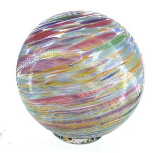 May include: A spherical glass paperweight featuring a swirling pattern of multicolored streaks in shades of pink, purple, yellow, green, and blue. The clear glass surface has a smooth, polished finish that reflects light across the vibrant colors.