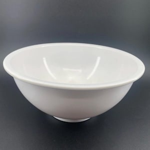 May include: A white mixing bowl with a rounded shape and a slightly flared rim. The bowl is made of a solid, opaque material and sits on a dark surface. The bowl is designed for food preparation and mixing.