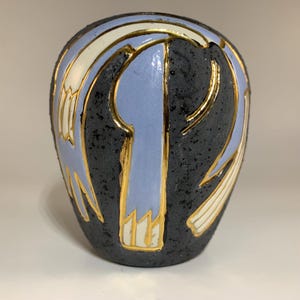 Puede incluir: Un pequeño jarrón de cerámica negra con una superficie texturizada. El jarrón está decorado con diseños geométricos blancos y dorados y un esmalte azul claro.