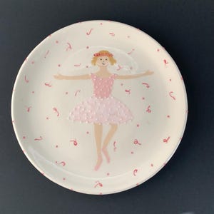 Könnte beinhalten: Ein weißer Keramikteller mit einer handbemalten rosa Ballerina in einem weißen Tutu. Die Ballerina ist von rosa Punkten und Wirbeln umgeben.