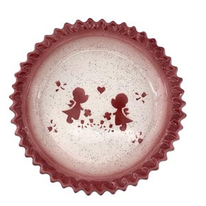 Puede incluir: Un molde redondo de cerámica para tarta con borde ondulado y festoneado. El interior presenta una base crema moteada con un borde en degradado rojo oscuro. El centro muestra un gráfico de silueta en rojo oscuro de dos figuras frente a frente.