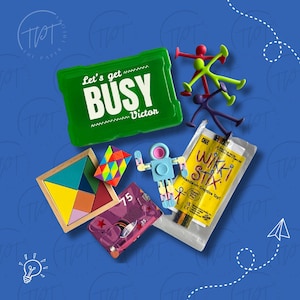 May include: A collection of colorful toys and games on a blue background. Includes a green container with the text "Let's get BUSY Victor", a wooden tangram puzzle, a Rubik's cube, and a package of Wikki Stix.