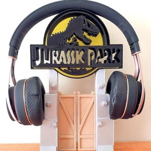Könnte beinhalten: Ein 3D-gedruckter Kopfhörerständer mit dem Jurassic Park-Logo. Der Ständer ist grau und hat ein braunes Tor. Das Logo ist schwarz und gelb mit einer Dinosauriersilhouette.