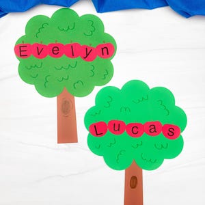May include: Two handmade paper tree cutouts. The trees have green, wavy-edged leaves and brown trunks. Red apple cutouts with the names "Evelyn" and "Lucas" are attached to the leaves. A blue fabric is in the background.