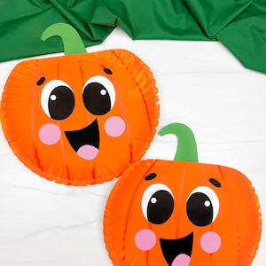 Può includere: Due zucche di piatti di carta arancioni con facce sorridenti, occhi neri, guance rosa e gambi verdi. Le zucche sono su una superficie bianca con uno sfondo di tessuto verde, perfette per le decorazioni di Halloween.