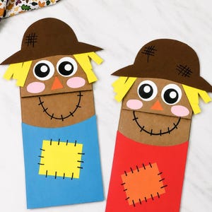 Peut inclure: Deux marionnettes en sac en papier en forme d'épouvantails, chacune dotée d'un chapeau marron avec des hachures noires, des cheveux en papier jaune, de grands yeux blancs et noirs, un nez orange triangulaire, des joues roses et une bouche cousue.