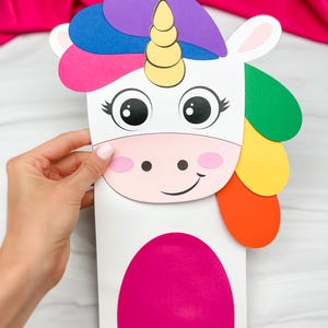 Op de afbeelding: Een kleurrijk papieren eenhoorn knutselwerk met een witte body, roze wangen en een regenboog manen. De eenhoorn heeft grote ogen en een gouden hoorn. Het knutselwerk wordt door een hand vastgehouden tegen een roze achtergrond.
