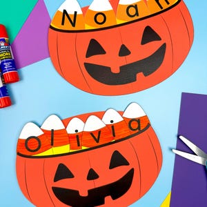 May include: Two orange paper pumpkin cutouts with black jack-o'-lantern faces. Each pumpkin has candy corn with a name on top. One says "Noah" and the other says "Olivia". The background is blue, purple, and yellow.