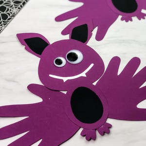 May include: Two purple bat-shaped crafts made from construction paper, with googly eyes and black accents. The bats have hand-shaped wings and a black oval body. A black and white spiderweb pattern is visible in the background.