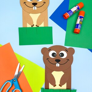 May include: Two paper groundhog puppets with smiling faces and a green grass base. Craft supplies, including glue sticks, scissors, and colorful paper, are arranged around the puppets on a light blue background. A fun craft project.