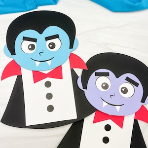 Könnte beinhalten: Zwei Vampir-Bastelarbeiten aus Papier mit schwarzem Haar, roten Kragen, weißen Hemden mit drei schwarzen Knöpfen und roten Fliegen. Ein Vampir hat ein hellblaues Gesicht, der andere ein hellviolettes, beide mit weißen Fangzähnen.