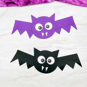 May include: Two paper bat crafts, one purple and one black, each featuring a circular body, pointed ears, and jagged wings. Both bats have large white eyes with black pupils and two small white fangs. The text simpleeverydaymom.com is visible.
