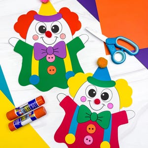 May include: Two paper clown puppets featuring colorful construction paper, pom-pom noses, and button details. One puppet wears a green outfit with a purple bow tie, while the other wears a red outfit with a green bow tie. Scissors and two glue sticks are present.