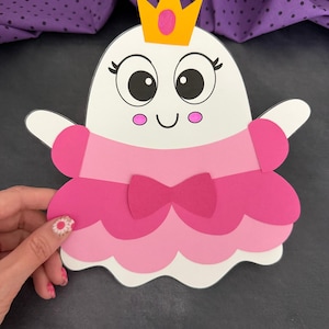 Op de afbeelding: Een witte geest-uitsparing die een roze jurk draagt met een geschulpte zoom en een donkerroze strik in de taille. De geest heeft grote zwarte ogen met lange wimpers, roze wangen en een gele kroon met een roze detail in het midden.