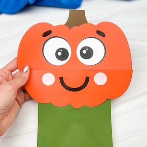 May include: A paper craft pumpkin puppet featuring a bright orange pumpkin head with a brown stem, large white eyes with black pupils, black eyebrows, a black smile, and pink cheeks, attached to a rectangular green paper body base.