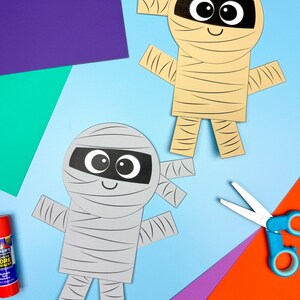 May include: Two paper mummy crafts with black eye details and wrapped bandages. One mummy is beige, the other is grey. Craft supplies, including scissors, glue, and colourful paper, are arranged on a blue background.