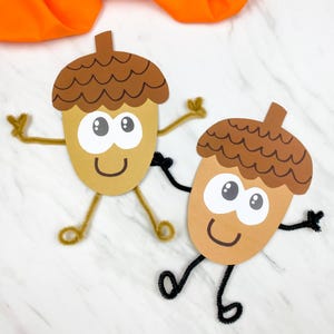 May include: Two paper acorn crafts with brown caps and tan bodies. Each acorn features large white eyes with black pupils, a drawn smile, and pipe cleaner arms and legs. One acorn has gold pipe cleaners, while the other has black pipe cleaners.