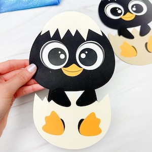 May include: Two penguin cutouts, one held by a hand, featuring a black body, white eyes, and a yellow beak. The penguin is emerging from a cream-colored eggshell with orange feet. A second penguin cutout is visible in the background.