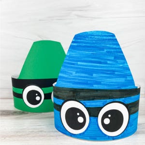 May include: Two paper crowns shaped like crayons, one in bright green and one in vibrant blue. Each crown features a wide black band around the base with two large, cartoon-style white eyes with black pupils centered on the front of the band.
