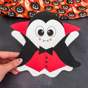 Könnte beinhalten: Ein Papier-Ausschnitt eines Cartoon-Geistes, der als Vampir verkleidet ist. Der Geist hat ein weißes Gesicht, große schwarze Augen, rosa Wangen und zwei kleine Fangzähne. Er trägt einen schwarzen Umhang mit rotem Futter, eine schwarze Fliege und eine rote Weste.