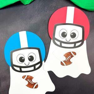May include: Two paper cutouts with a football theme. One has a blue helmet, the other a red helmet, both with a white stripe. Each cutout has a smiling face and holds a brown football. The background is dark grey with green fabric at the top.