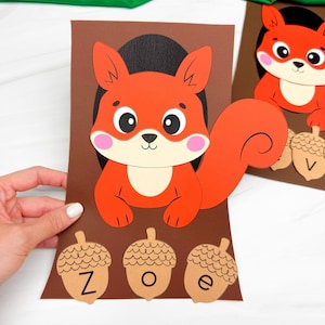May include: A brown paper craft featuring a cartoon squirrel with orange fur, a cream-colored belly, and pink cheeks. Below the squirrel are three tan paper acorns, each labeled with the black letters Z, O, and e, arranged horizontally.
