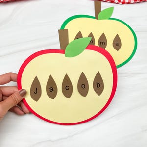 May include: Two apple-shaped paper crafts, one red and one green, each with a yellow center. The red apple has the name "Jacob" spelled out with brown seed shapes. The green apple has the name "Anna" spelled out. Both have brown stems and green leaves.