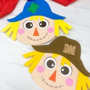 May include: Two paper scarecrow faces with yellow paper hair, orange triangular noses, pink circular cheeks, and black stitched mouths. One scarecrow wears a blue hat with a green patch, and the other wears a brown hat with a tan patch.