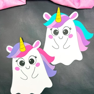 May include: Two whimsical white paper cutouts of unicorn ghosts with colourful manes, pink cheeks, and yellow horns. The background is a dark grey surface with a pink fabric in the upper left corner.