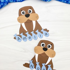 Puede incluir: Dos recortes de morsa marrones con hocicos color canela y colmillos blancos. Cada morsa tiene un nombre escrito en etiquetas azules con forma de pez. Una dice "Amelia" y la otra "Noah". Un tejido azul con copos de nieve está en el fondo.