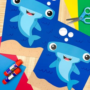 Könnte beinhalten: Zwei Papierbastelprojekte mit einem blauen Hammerhai auf dunkelblauem Hintergrund mit beigefarbenem Sand am Boden und weißen Blasen. Der Hai hat große Augen, ein breites Maul mit weißen Zähnen und Details am hellblauen Unterbauch.