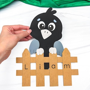 Puede incluir: Un recorte de papel de un pájaro negro con alas azules asomándose por encima de una valla de madera clara. La valla tiene el nombre Liam impreso en letras negras sobre los listones verticales. El pájaro tiene ojos grandes y pico blancos.